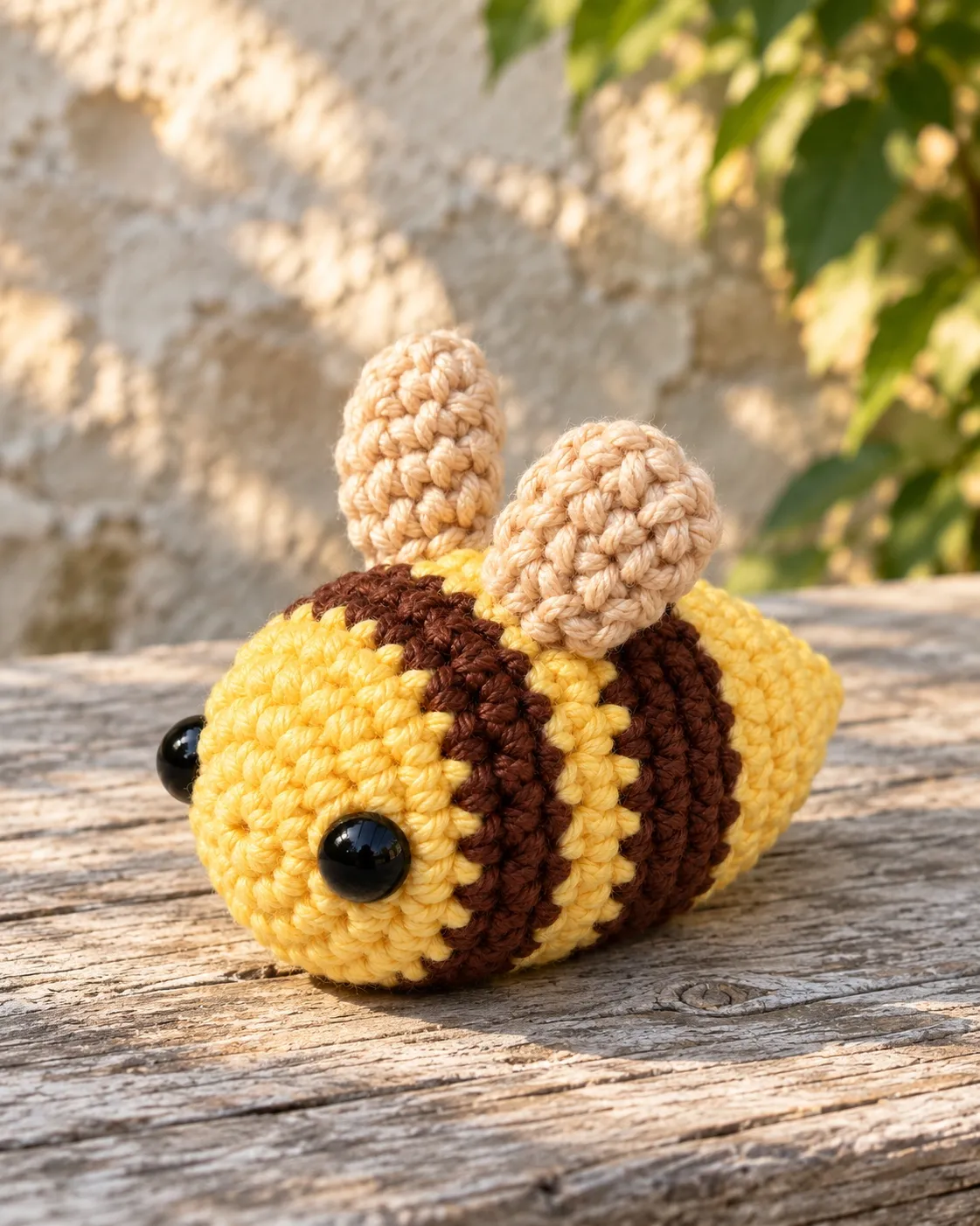 Illustration: a butter-yellow crocheted bee with two brown stripes lying on its belly on a weathered wooden plank in front of a sunlit stone wall and green leaves, seen low from the front-left, with two glossy black eyes on its round face and two cream wings standing on its back.