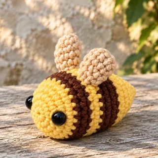 Illustration: a butter-yellow crocheted bee with two brown stripes lying on its belly on a weathered wooden plank in front of a sunlit stone wall and green leaves, seen low from the front-left, with two glossy black eyes on its round face and two cream wings standing on its back.