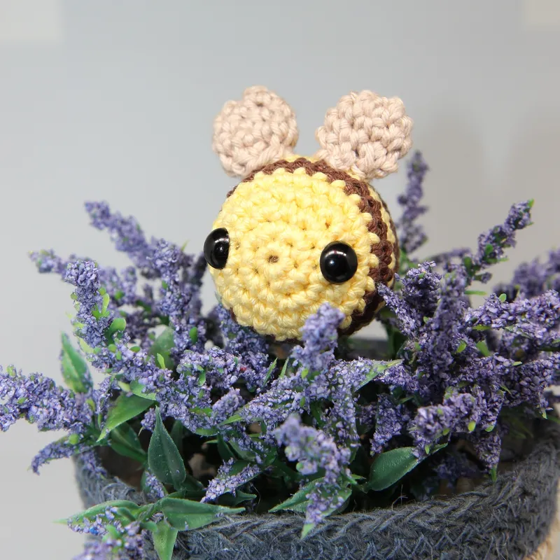 Photograph: the finished bee peeking out of a pot of lavender, its round yellow face with two black eyes, a brown stripe at the edge and two beige wings standing up behind.
