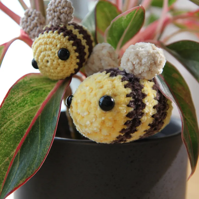 Photograph: two bees in the leaves of a potted plant, the larger one in soft yellow plush yarn with fluffy cream wings in front, the smaller cotton bee above it.