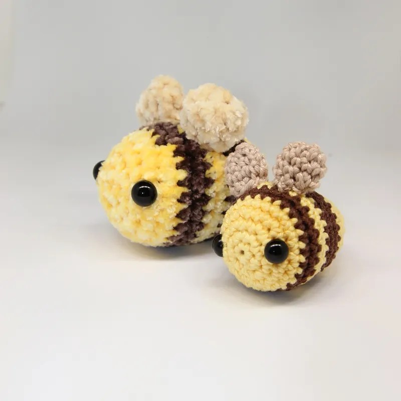 Photograph: the plush bee and the smaller cotton bee side by side on a white surface, both with black eyes, two brown stripes and two standing wings.