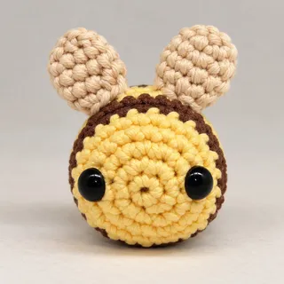 A small crocheted bee in butter yellow with two brown stripes, seen from the front, with two glossy black eyes on its round face and two cream wings standing on its back.