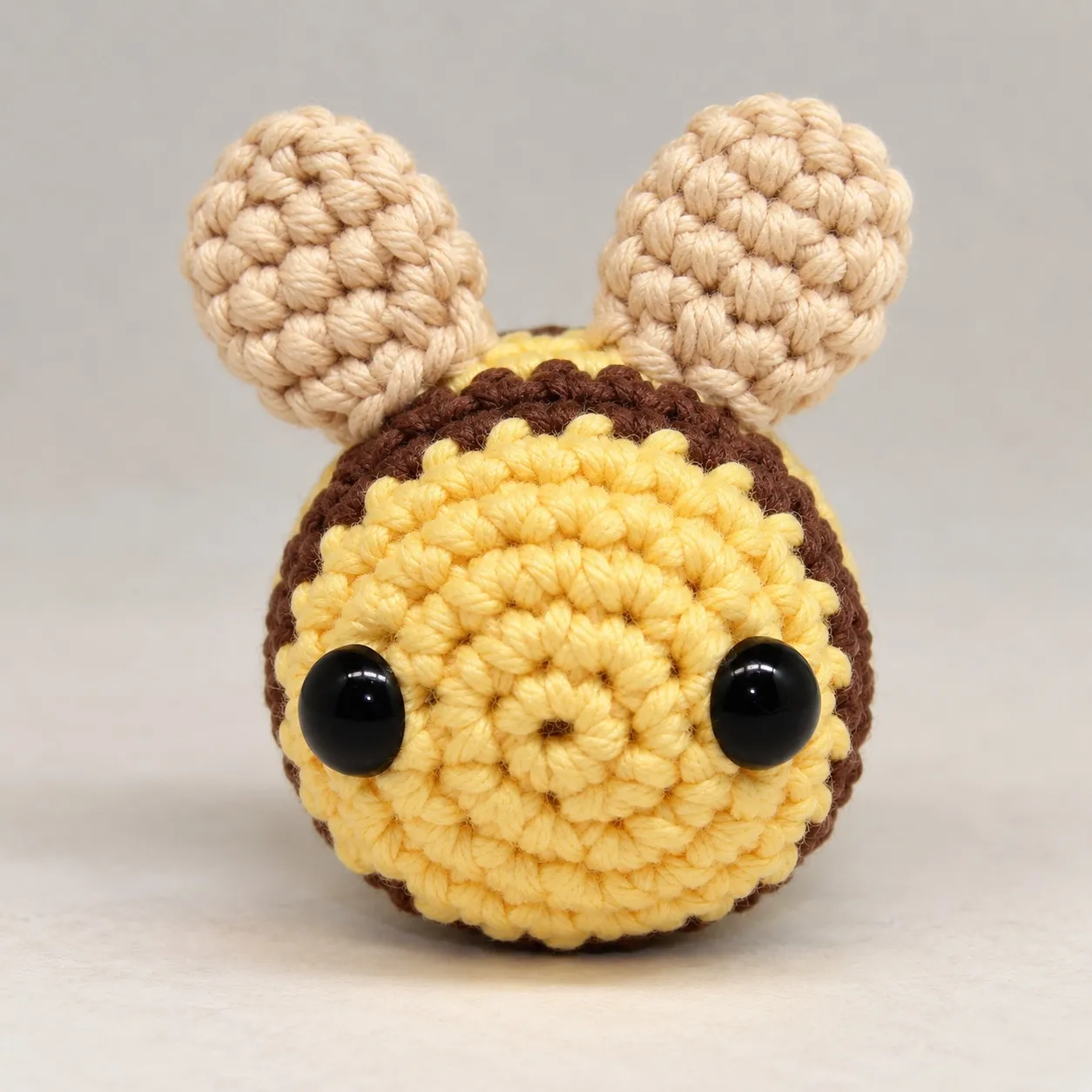 A small crocheted bee in butter yellow with two brown stripes, seen from the front, with two glossy black eyes on its round face and two cream wings standing on its back.
