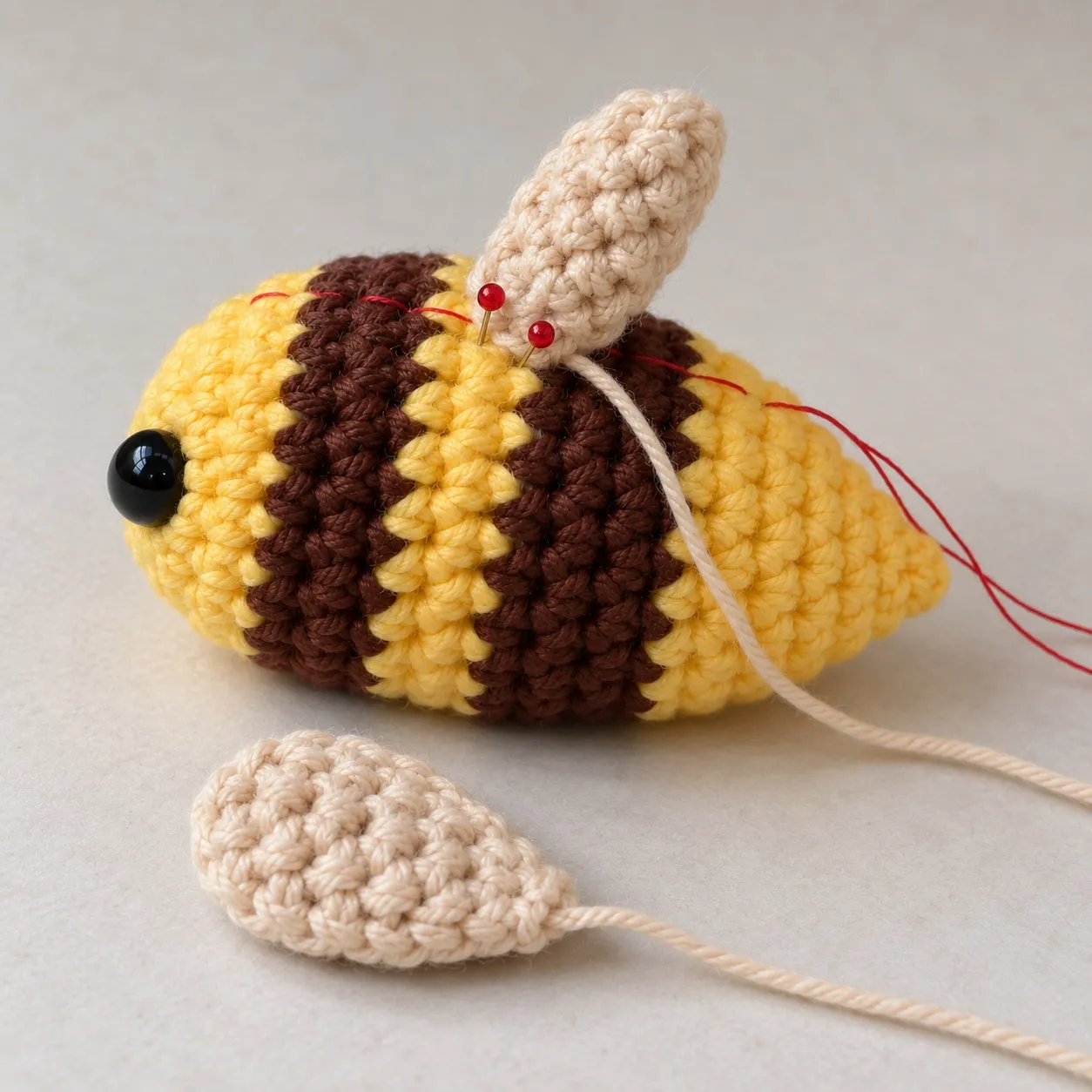 The bee from the side with one cream wing pinned upright on its back with two pins, a red basting thread along its back, and a second wing lying loose in front.