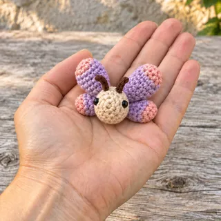 The small crocheted butterfly resting in the middle of an adult's open palm, a wooden plank and a sunlit stone wall out of focus behind.