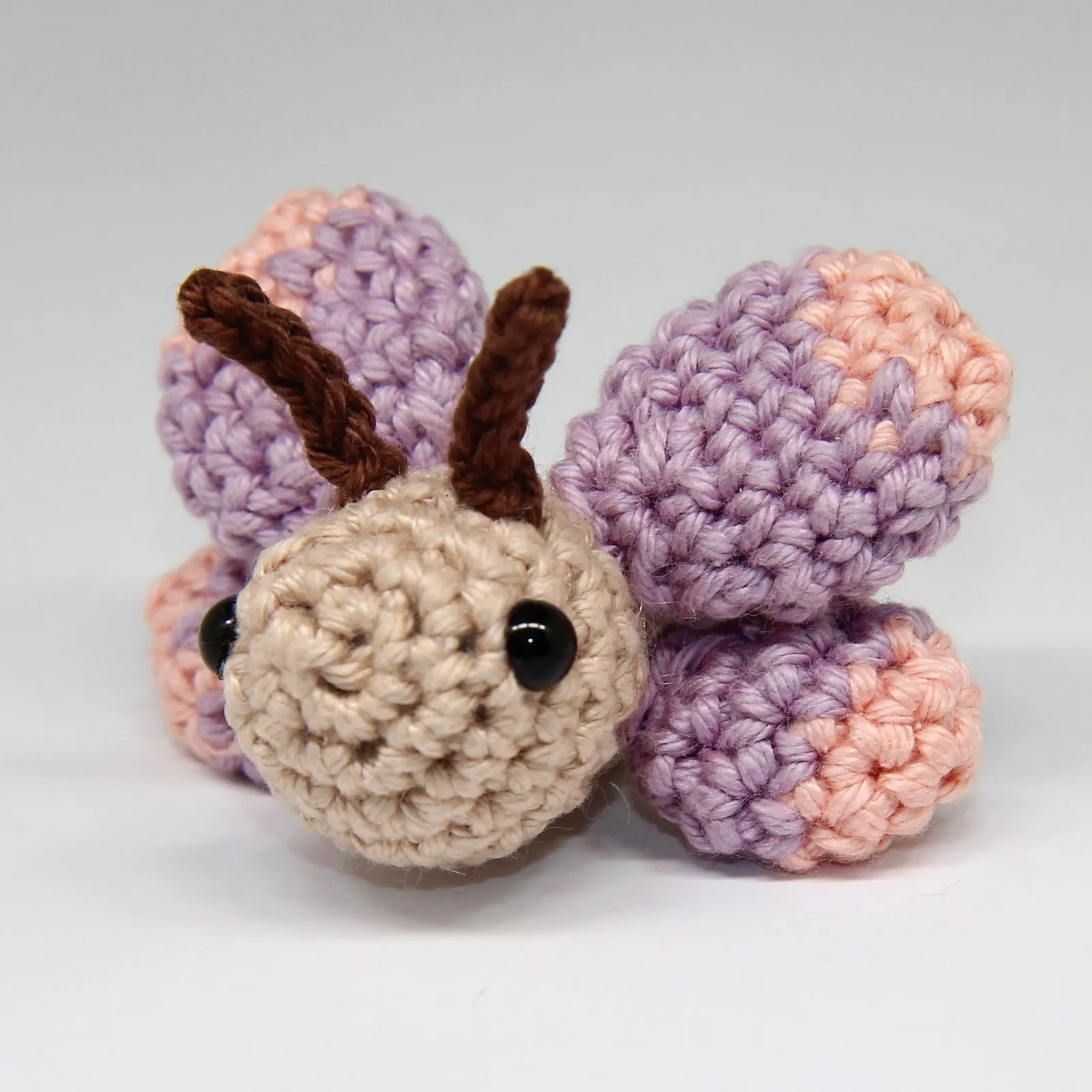 Photograph: the crocheted butterfly on a plain pale ground, turned slightly to one side, its beige head with two black eyes and two brown cord antennae between four lilac wings tipped in peach.
