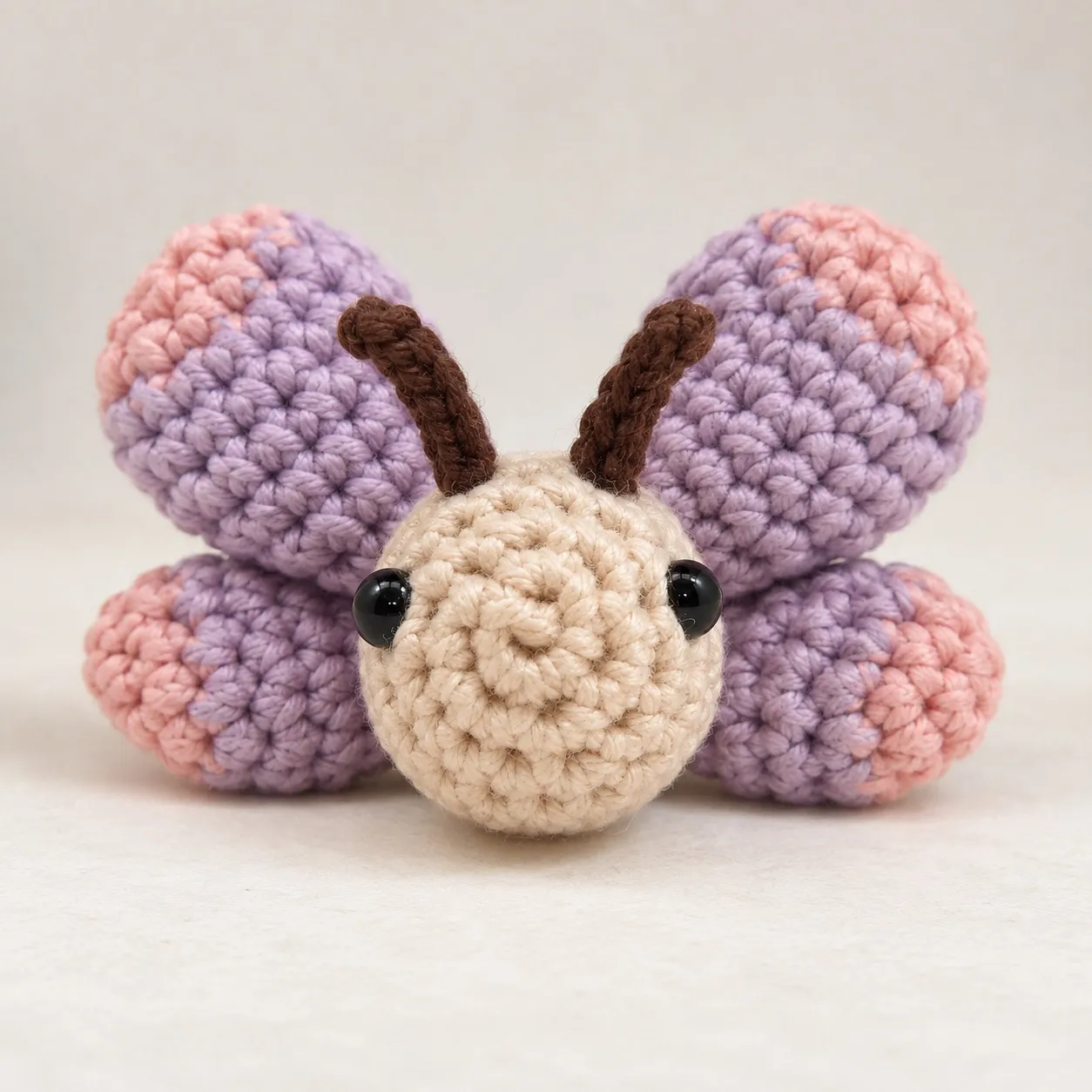 A small crocheted butterfly lying on its belly, seen from the front: a cream head with two glossy black eyes and two thin brown antennae, two lavender forewings standing up behind it and two smaller hindwings lying low at its sides, every wing tipped in dusty rose.