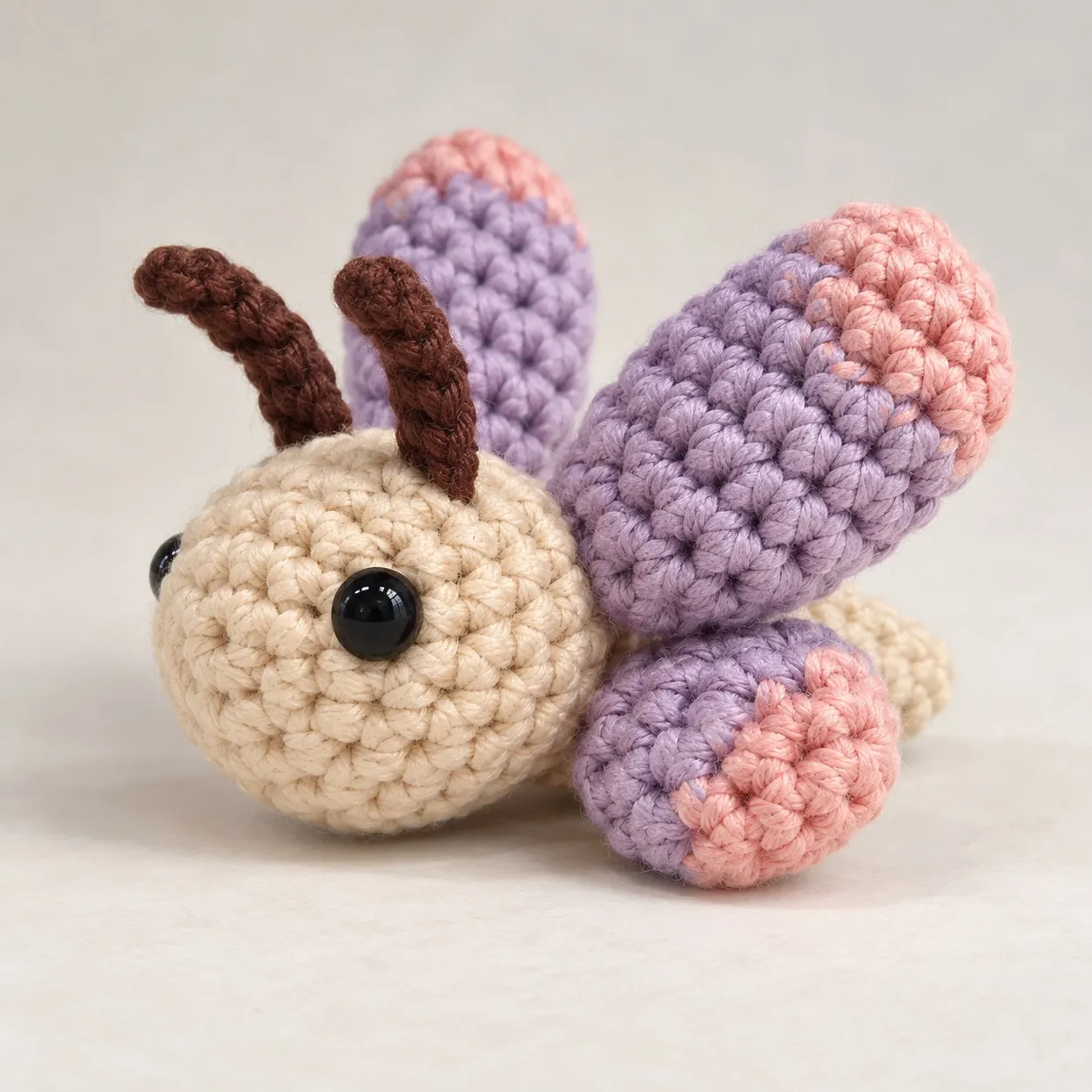 The crocheted butterfly from the front-left: the cream head with its eyes and thin brown antennae, two lavender forewings standing up behind it and a hindwing lying low at its side, every wing tipped in dusty rose.