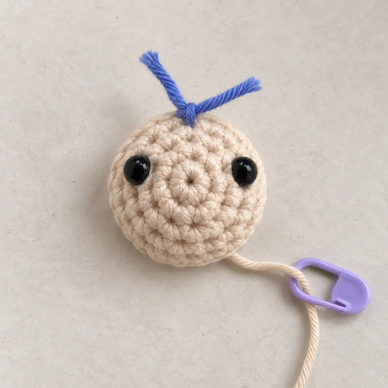 A shallow cream crocheted dome seen from above, with two black safety eyes pushed into it, a blue scrap of yarn tied at its top between them and the working loop held on a stitch marker.