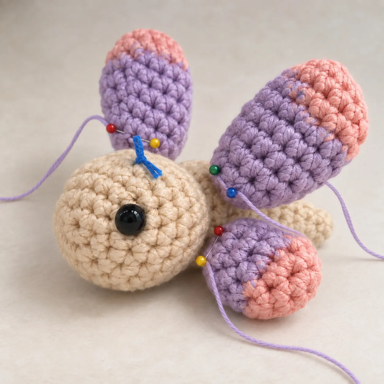 The closed cream body with four lavender wings pinned to it, two glass-headed pins at each wing's base, the upright pair above the low pair, a lavender tail from each wing and a blue scrap of yarn on top of the head.