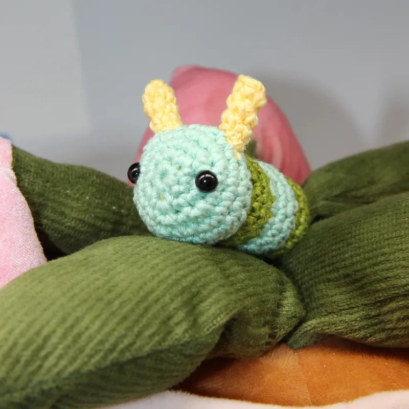 Photograph: the caterpillar lying on the green fabric leaves of a soft flower, its mint head turned toward the camera, the yellow antennae up and the green and mint bands of its body behind.