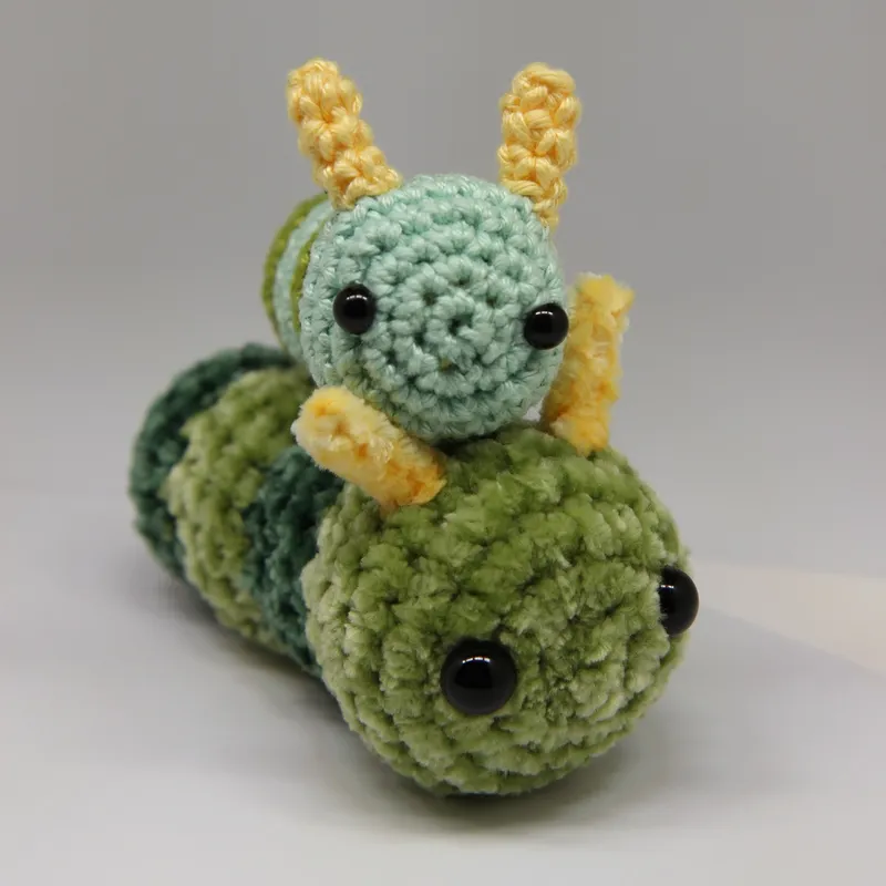Photograph: the cotton caterpillar resting on top of a much larger caterpillar in soft green plush yarn with darker green bands, both with black eyes and yellow antennae.