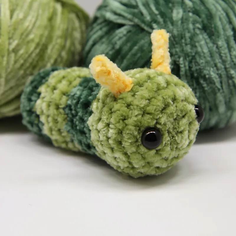 Photograph: the plush caterpillar in light and dark green bands lying in front of two balls of the same green plush yarn, its yellow antennae up.