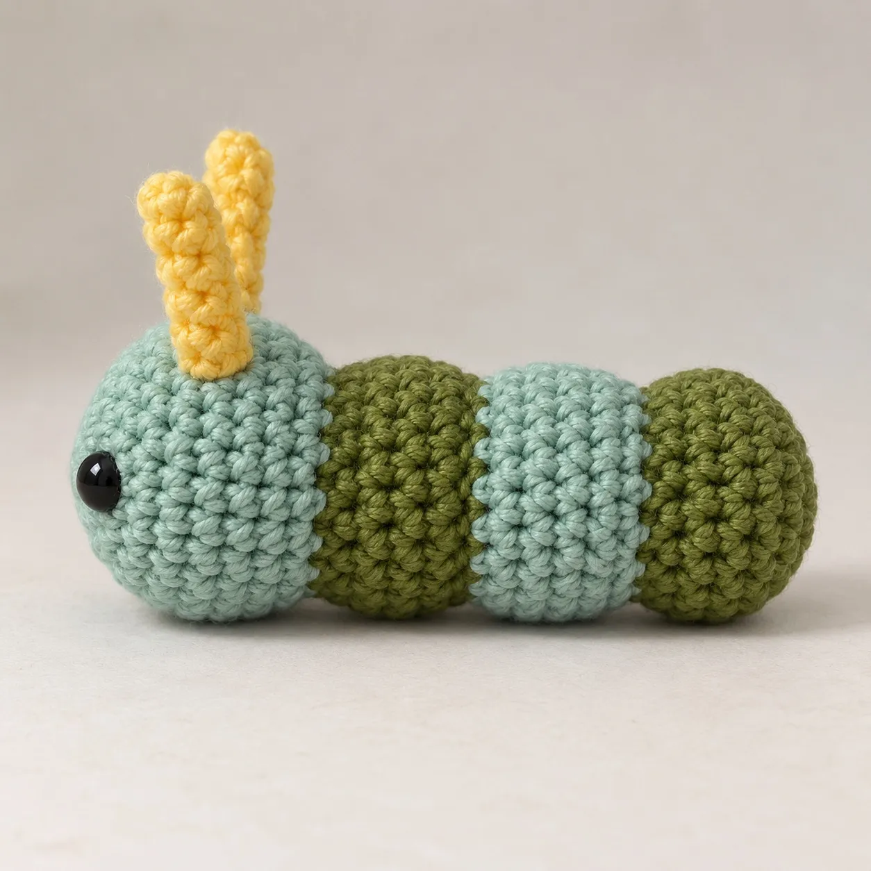 The same caterpillar from its left side: the sage-teal head with one eye and two yellow antennae, then three rounded bulges in alternating greens joined by shallow grooves.