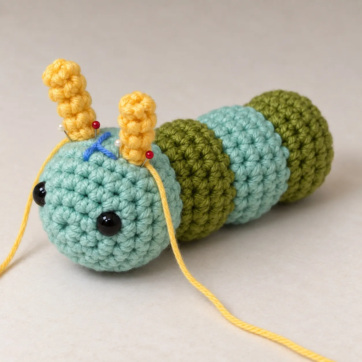 The caterpillar with two yellow crocheted antennae pinned through their bases to its head, their yarn tails trailing away, a blue yarn scrap tied between them.