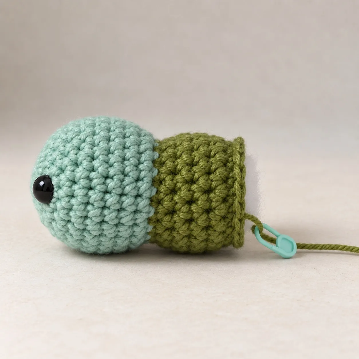 From the side, a sage-teal crocheted head with one eye, a shallow groove, and a moss-green bulge filled firm and still open at its far end, the working yarn on a stitch marker.
