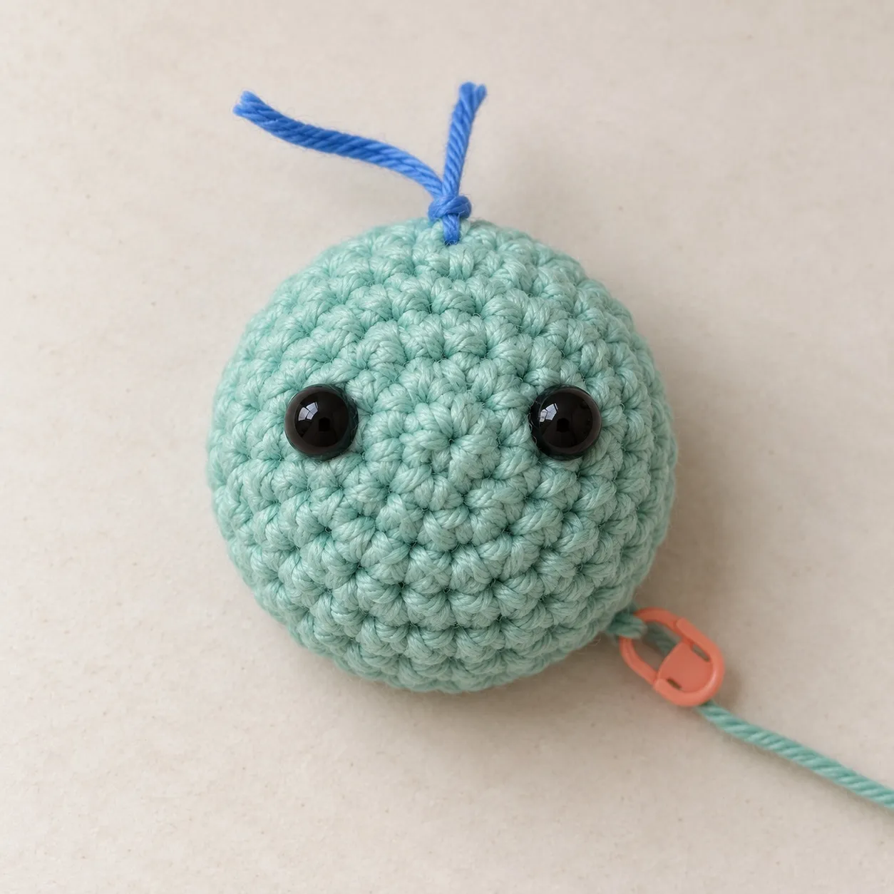 Seen from above, a small sage-teal crocheted dome with two black safety eyes, a blue yarn scrap tied at its top edge and the working yarn held on a stitch marker.