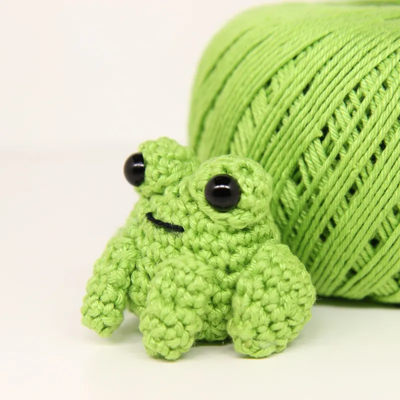 Photograph: the finished frog in bright green mercerized cotton, sitting on a white surface with its ball of yarn behind it, its two black eyes on raised cups and a straight stitched mouth.