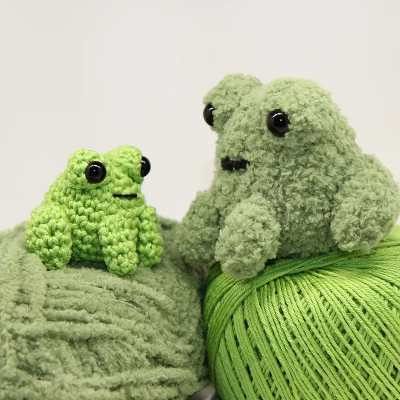 Photograph: the frog worked in sage green plush microfibre yarn, sitting on a ball of the same yarn, with the much smaller bright green cotton frog beside it on a second ball.
