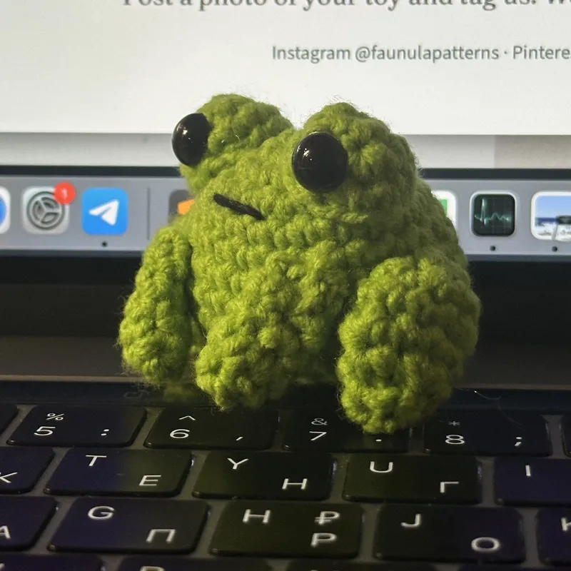 Photograph: a small green crocheted frog with large black eyes and a straight stitched mouth, sitting on a laptop keyboard.