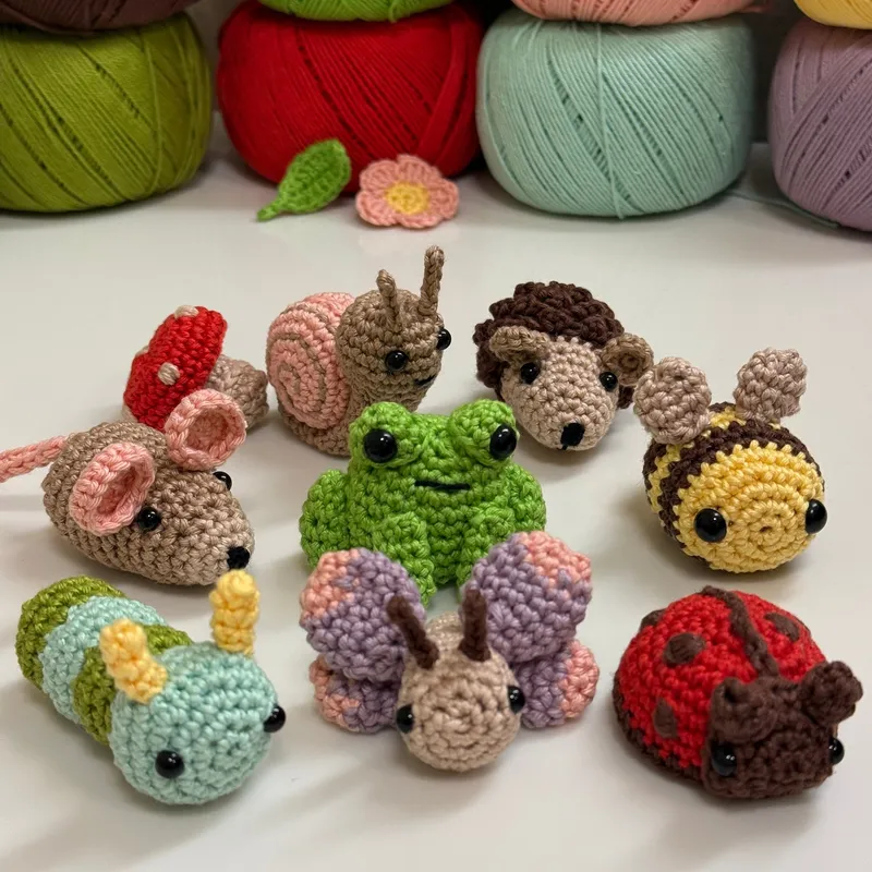 Photograph: the eight crocheted garden animals together in front of balls of cotton yarn — a hedgehog, snail, bee, frog, field mouse, butterfly, ladybug and caterpillar — with a red mushroom, a green leaf and a pink flower among them.