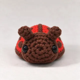 A small crocheted ladybug in brick red with a cocoa-brown head, two glossy black eyes and two short antennae, a brown line down its back and brown spots, seen from the front.