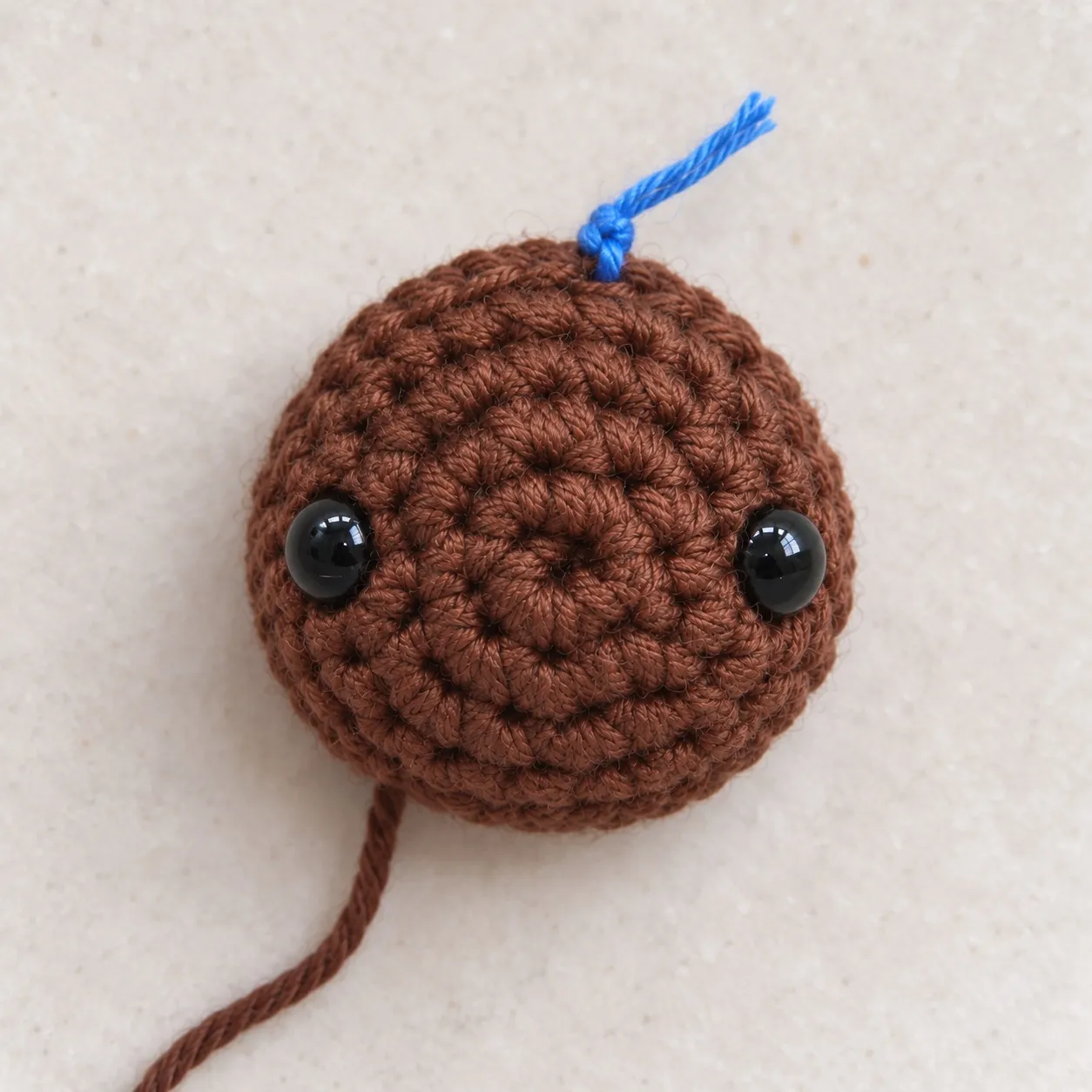 Seen from above, a small brown crocheted dome with two black safety eyes, a blue yarn scrap tied at its top edge and a long brown yarn tail.