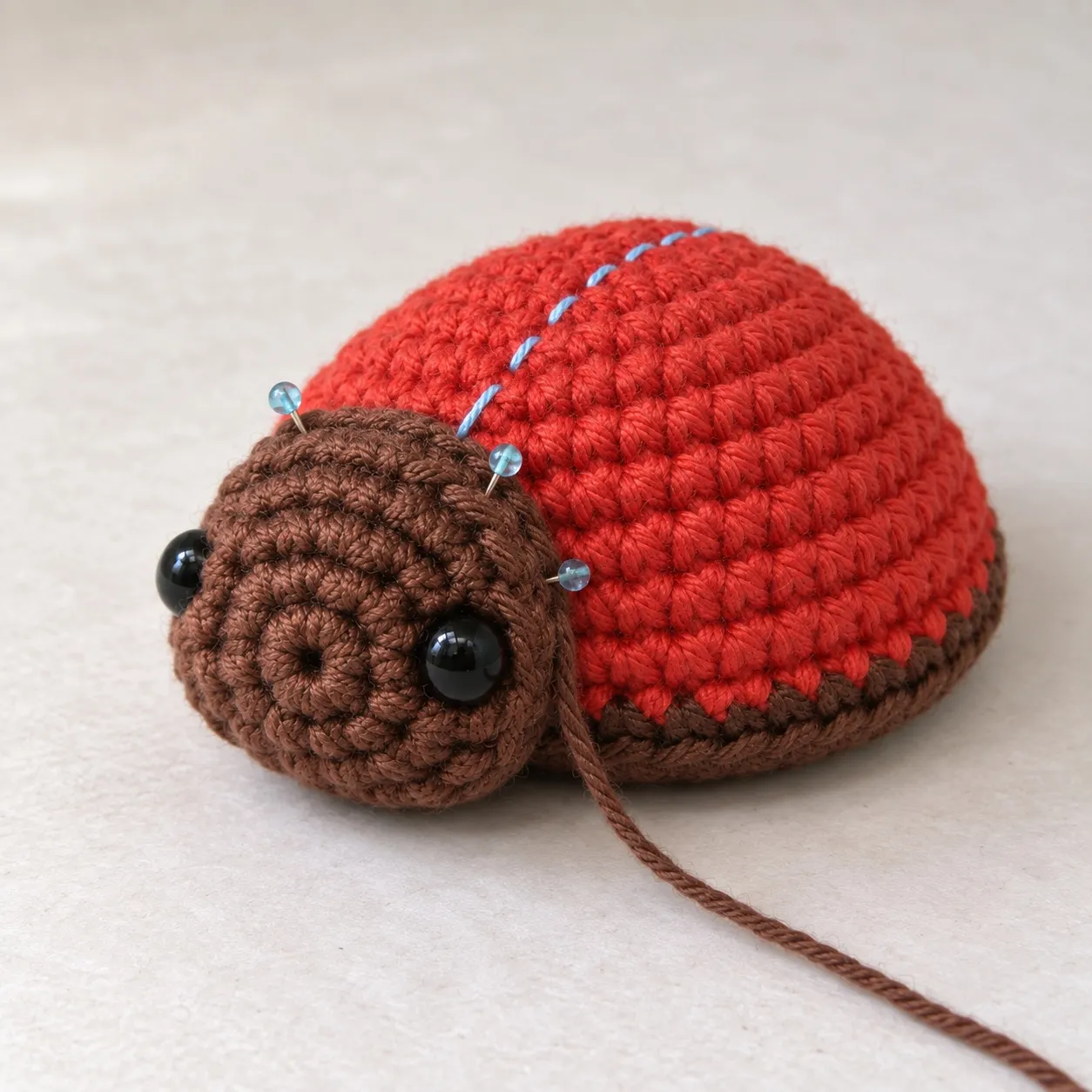 A plain red crocheted dome with a brown head pinned low to its front by pins through the head's edge, and a blue thread running over the top.