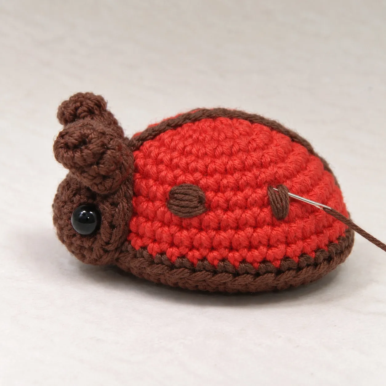 The ladybug from its left side with one brown embroidered spot finished and a second spot half laid in satin stitch, a threaded needle at its edge.